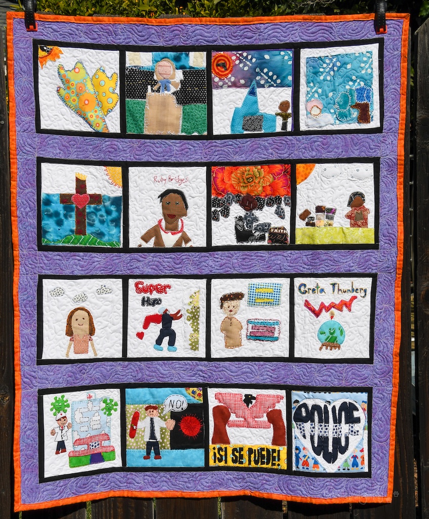 A quilt is shown hanging against a dark fence. The quilt has individual squares designed by students showing their personal hero activist (more recognizable ones include Super Deafy, Ruby Bridges, Greta Thurnberg, Cesar Chavez). The squares are sashed together with black fabric and framed in purple. A bright orange binding completes the quilt.