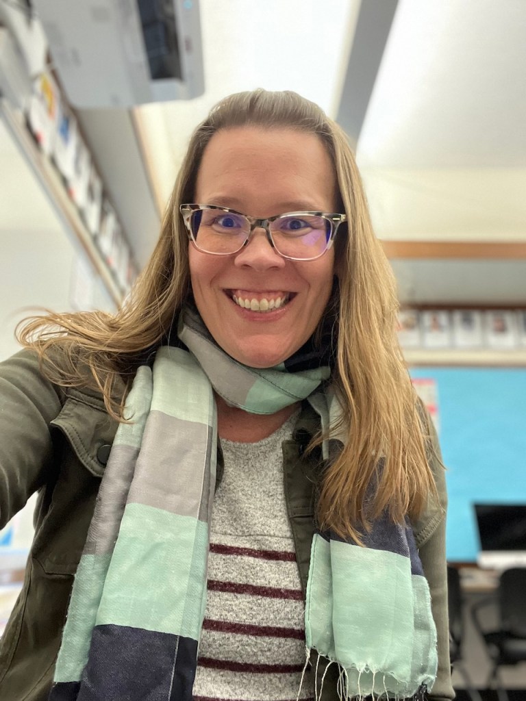 Keila, a white woman with glasses and long light brown hair, is pictured smiling with a gray sweater and light teal and gray scarf.