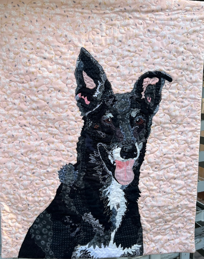 A collage of a dog hangs against an outdoor lattice background. The dog is black with one ear erect and the other flopped over slightly at the tip. The dog's mouth is open, with a pink tongue hanging out. The dog has white markings around it's nose, down it's neck, and across it's chest.