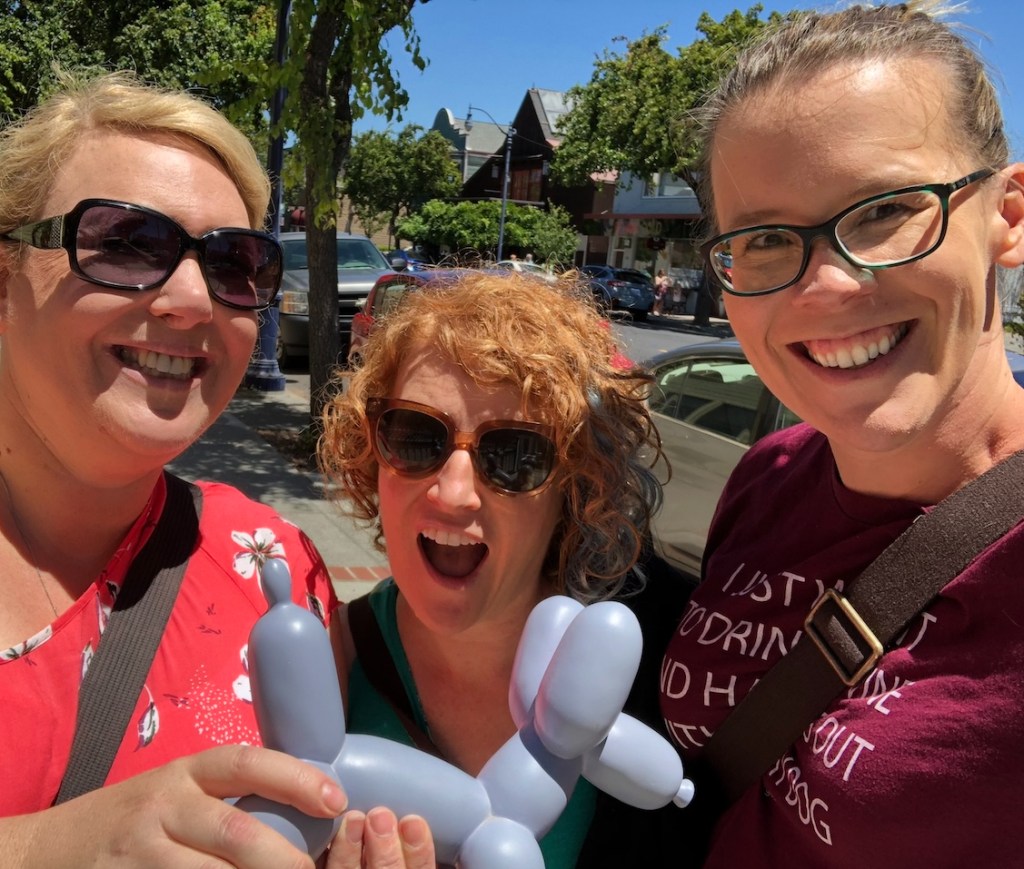 Three woman are pictured outside on a city street with parked cars and trees. The two on the left are holding a lavender balloon dog sculpture. The woman on the left is blonde with dark sunglasses. The woman in the center has curly red hair with a teal stripe and is also wearing sunglasses. The woman on the right has light brown hair pulled back and regular glasses. All three are smiling into the camera.