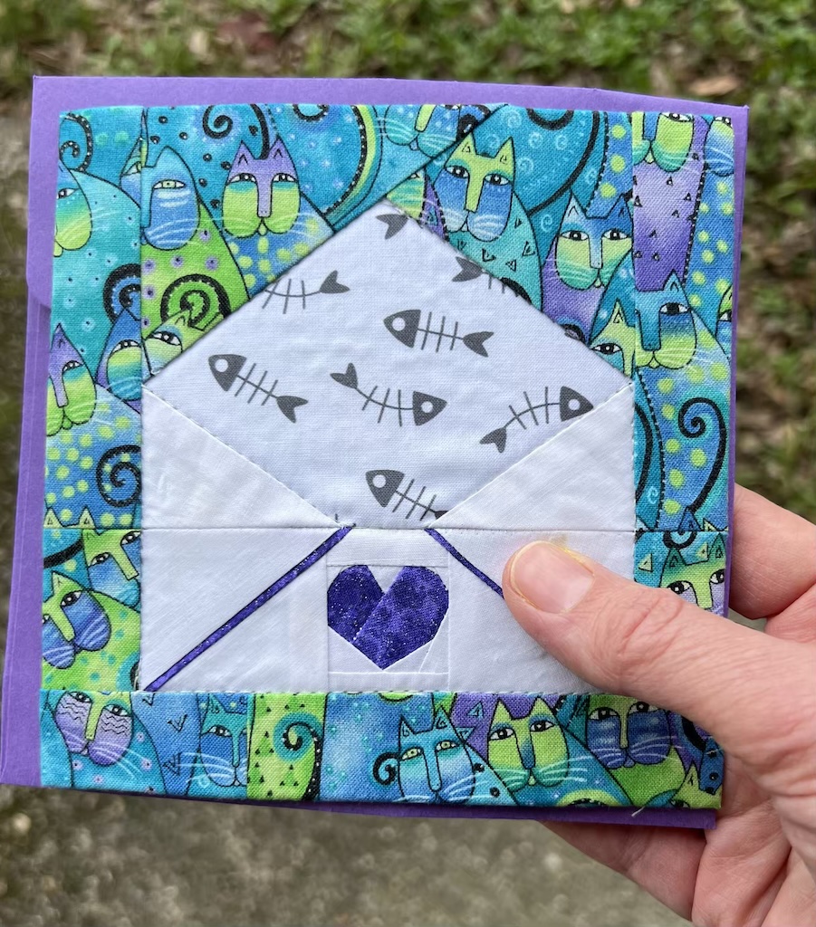 A white person's hand holds a purple and teal, square fabric greeting card. The background fabric is green, blue, and purple whimsical kitties. The foreground shows a foundation paper pieced white envelope with fish bones on the interior of the flap and a purple heart on the lower portion of the outside of the envelope.