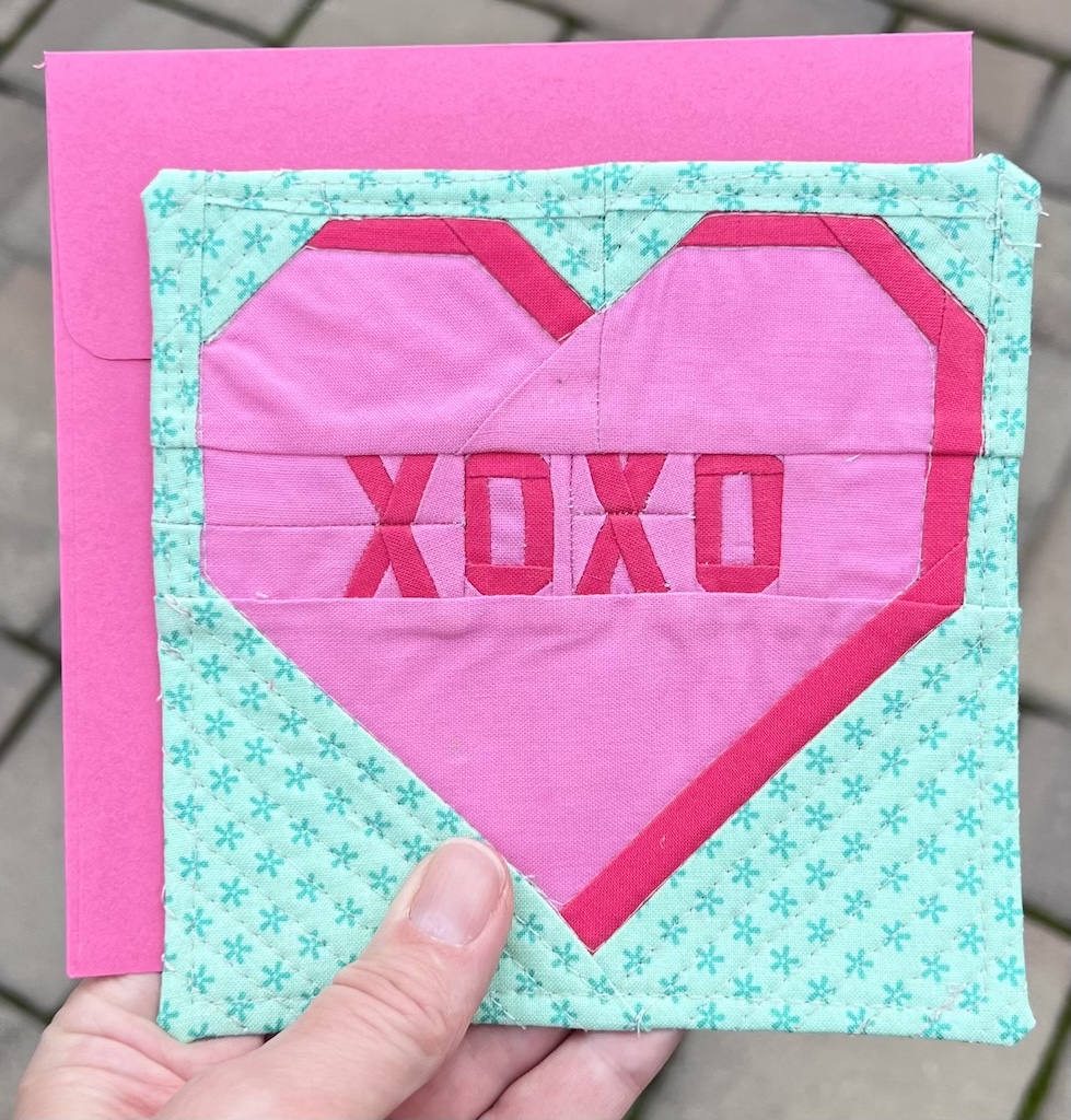 The hand of a white person holds a bright pink envelope with a green and pink fabric greeting card on top. In the background are gray paving stones. The greeting card has a bright pink conversation heart with darker pink "XOXO" in the middle and darker pink shading along the right side. The bright mint/teal background of the card has asterisks. The heart is echo quilted into the background.