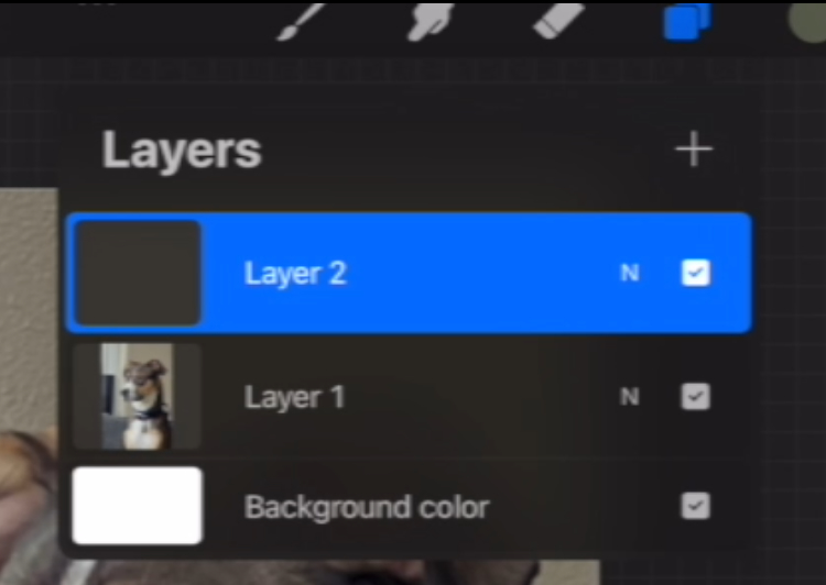 A screenshot of the Procreate app shows a brush, pointer, eraser, layerd squares, and a gray circle along the top menu bar. The double squares have been selected so are shaded blue. In the Layers menu, three layers are shown named Background color (with a white image), Layer 1 (with a photograph of a dog) and Layer 2 (gray image).