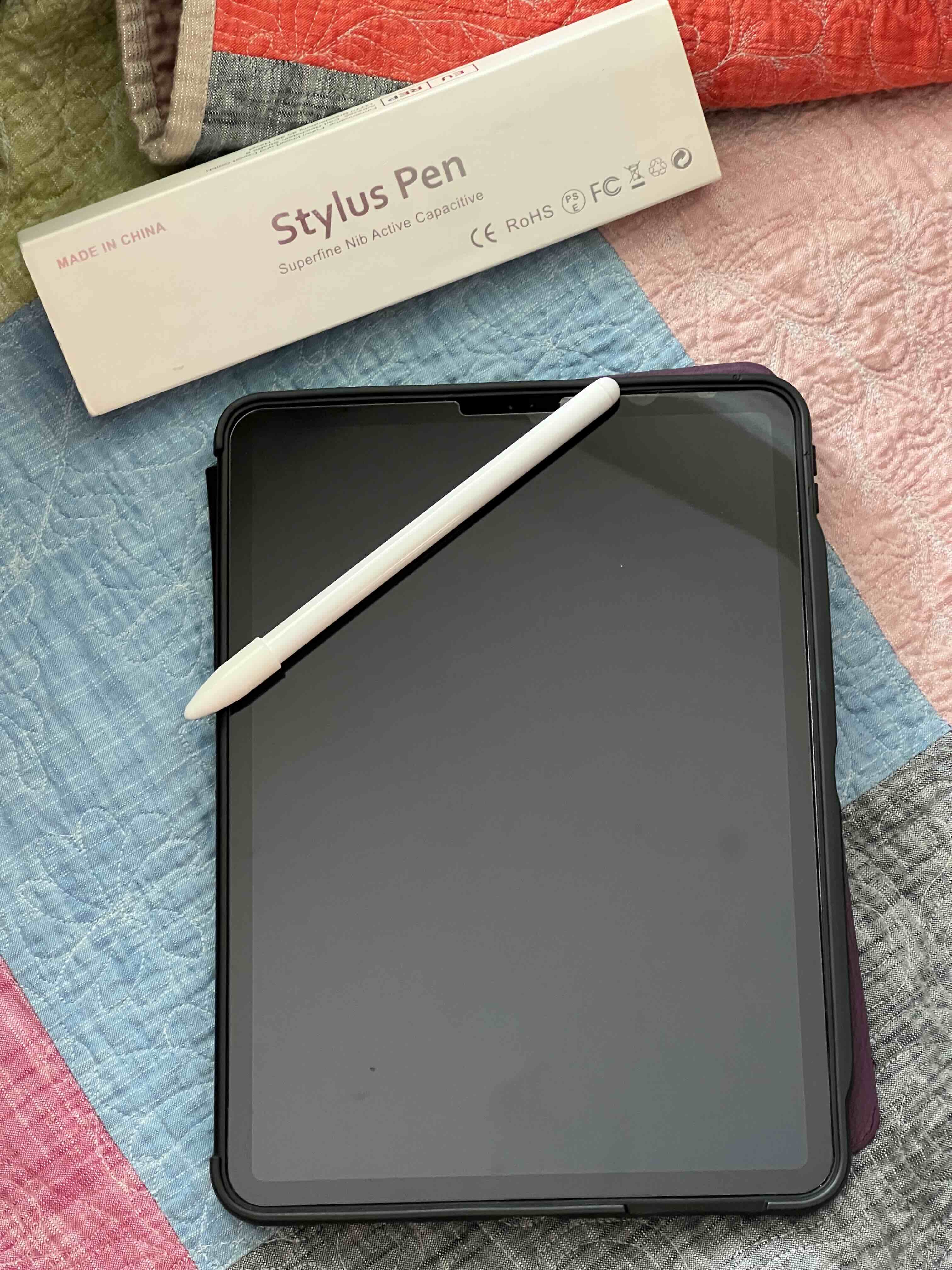 A black iPad and white stylus are pictured with the backdrop of a quilt with blue, pink, gray, orange, green, and rose colored squares. The stylus' original box is above the ipad and reads "Made in China. Stylus Pen. Superfine Nib Active Capacitive."