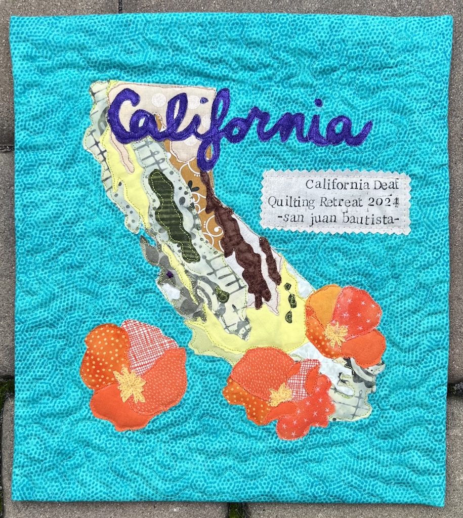 A small teal collage is pictured against some pavers. A topographical map of California is pictured in greens and browns. In purple cursive, "California" is across the top and three orange poppies are at the bottom of the collage. In slightly messy stamping you can read "California Deaf Quilting Retreat 2024 ~san juan bautista~