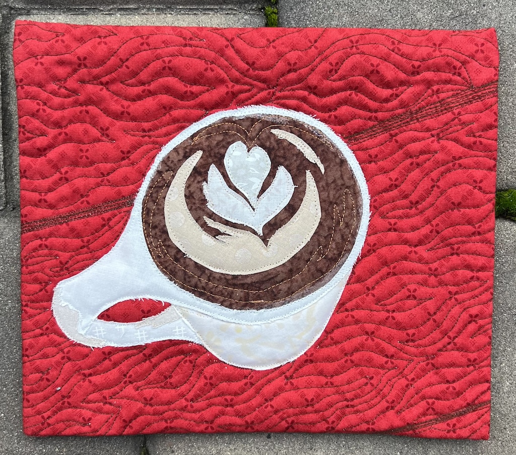 A fabric collage of a coffee cup is pictured with a bright red background. The mug is a white coffee mug with latte art (a heart) on the top. In the background is stitching intended to look like woodgrain planks of a table.