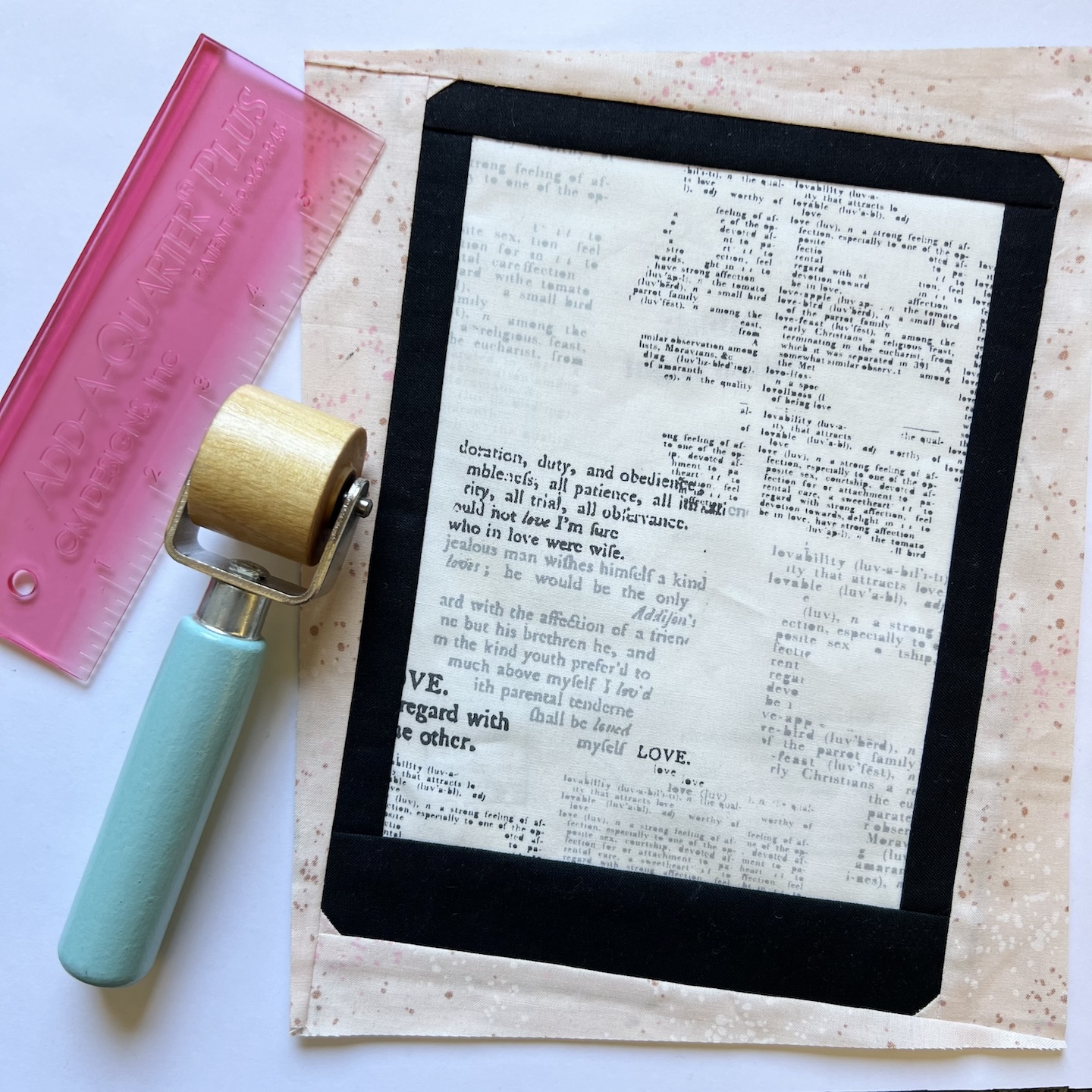 A fabric block is shown that depicts a black, non branded tablet with multiple lines of text on the center fabric "screen". Peach, gold, and white flecked fabric frame the block. A pink Add a Quarter Plus ruler and teal/wood seam roller lie next to the block against a white background.