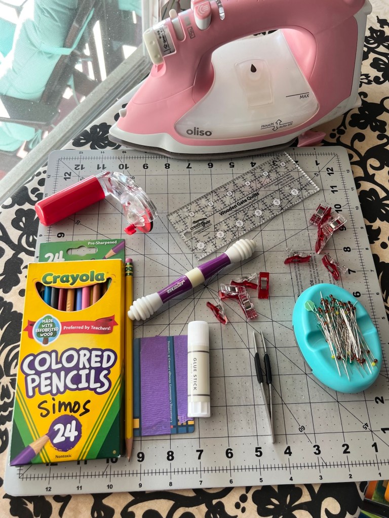 A small gray cutting mat rests on a black and white patterned ironing board. Pictured on top of it are a set of colored pencils, a number two pencil, a library card, a school glue stick, tweezers, sewing pins, sewing clips, a seam ripper, a small quilting ruler, and a rotary cutter. Behind it is a pink Oliso iron.,