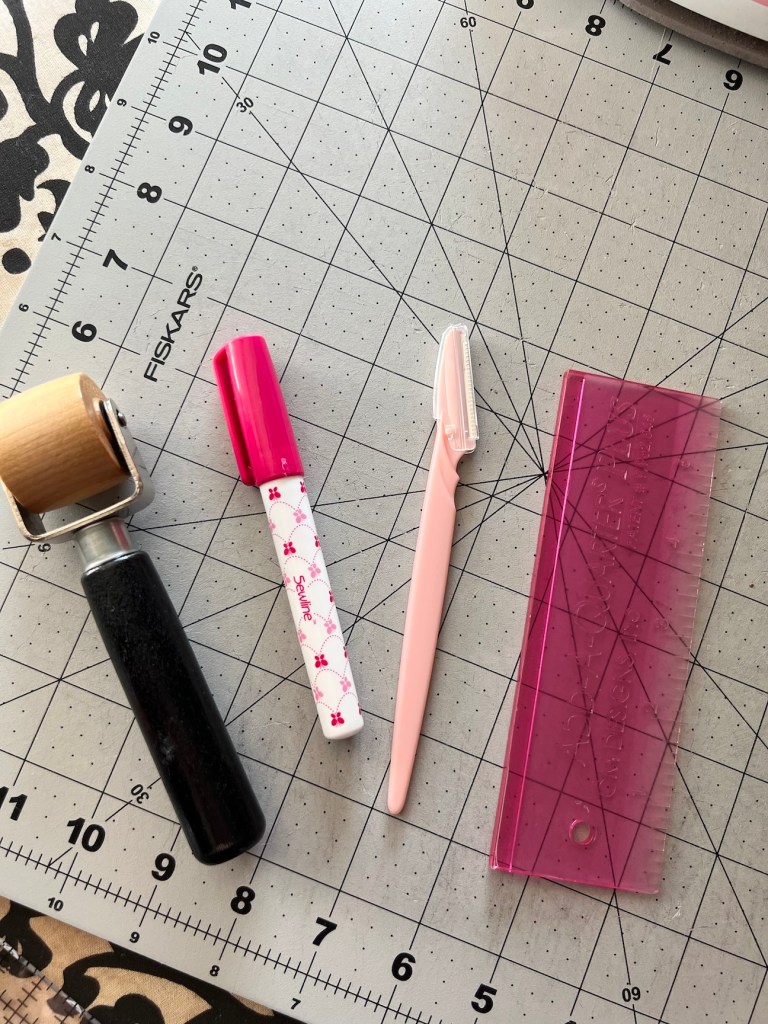 A small gray cutting mat rests on a black and white patterned ironing board. On top of it is a wooden seam roller, a Sewline glue pen, a light pink eyebrow razor, and a dark pink Add a Quarter Plus ruler.