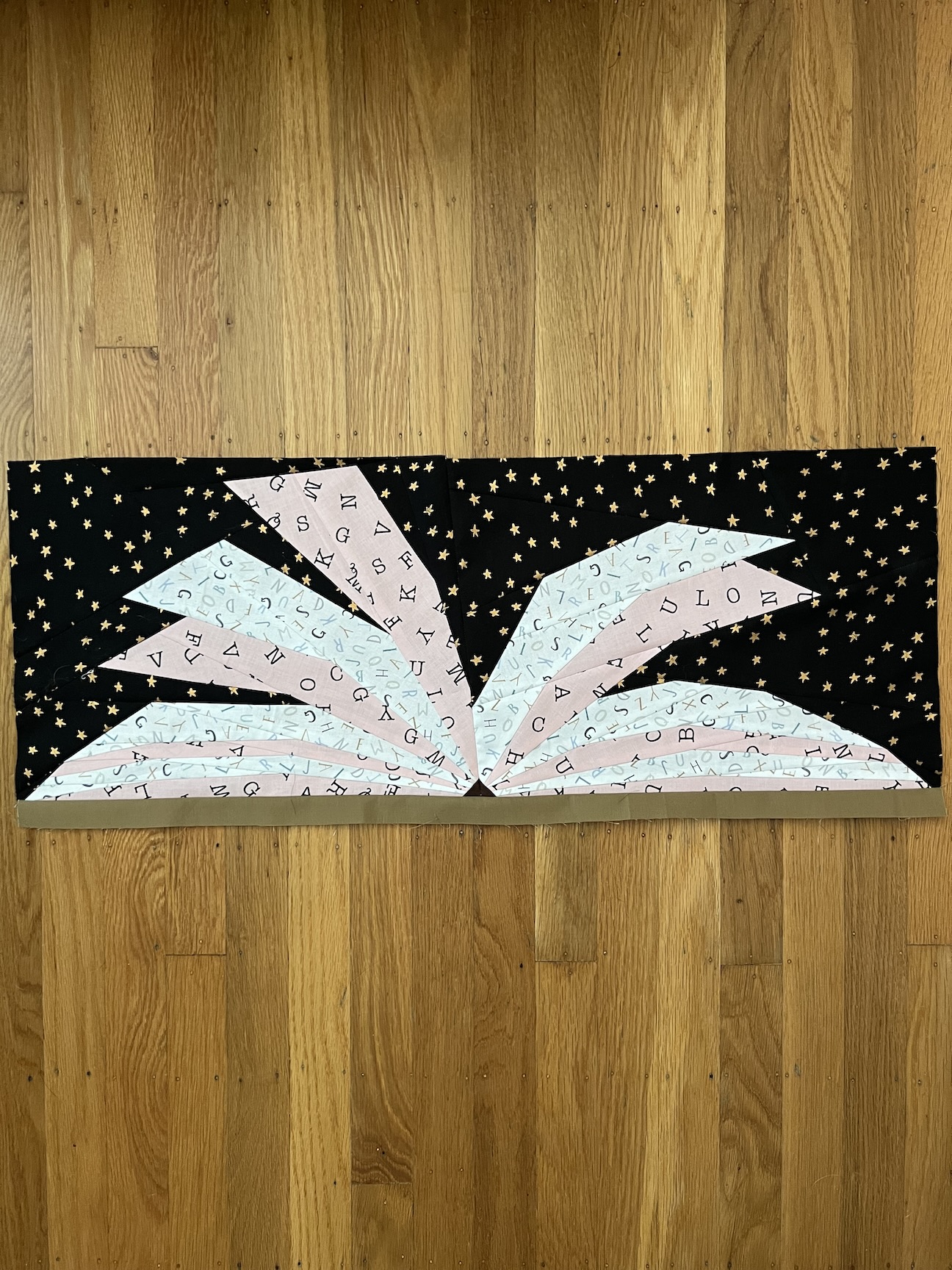 A quilt block is pictured lying on a hardwood floor with vertical boards. The block pictures an open book against a dark, starry background. The pages of the book alternate pink and white with alphabet letters, and the binding is two colors of brown.