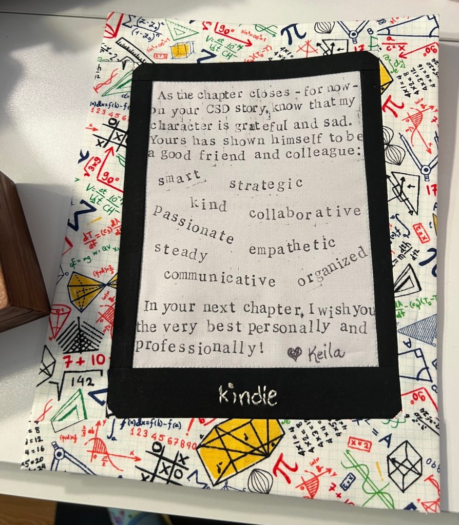 An individual quilt block is shown wrapped around a piece of cardstock to give it structure. In the background is fabric with math symbols. In the center is a representation of a kindle with a message to the recipient stamped messily in the center. The message reads: "As the chapter closes - for now - on your CSD story, know that my character is grateful and sad. Yours has shown himself to be a good friend and colleague: smart, strategic, kind, collaborative, passionate, empathetic, steady, communicative, organized. In your next chapter, I wish you the very best personally and professionally! (Heart shape) Keila