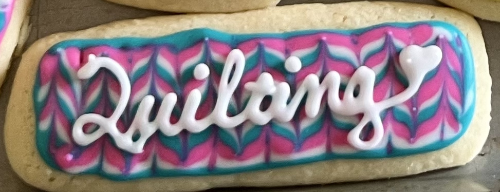 A sugar cookie is pictured with pink, white, and teal feathering. On top "Quilting" is written in white piped cursive with a heart at the end.