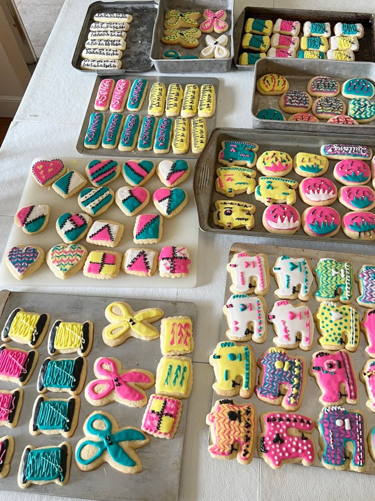 Brightly decorated sugar cookies (in teal, hot pink, yellow, and white are pictured drying on a table. There are cookies shaped like sewing machines, scissors, quilt blocks, rulers, and pincushions.