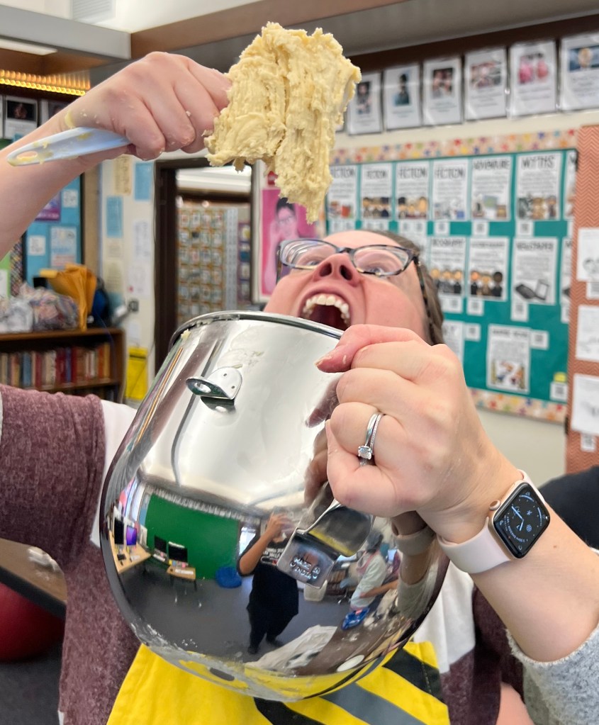 Keila (middle aged female in glasses and a rose and white striped shirt with a yellow apron) is pictured pretending to eat a huge scoop of cookie dough out of a mixing bowl she's holding. Her head is tipped back under the cookie dough which is shaggy and bumpy in appearance.