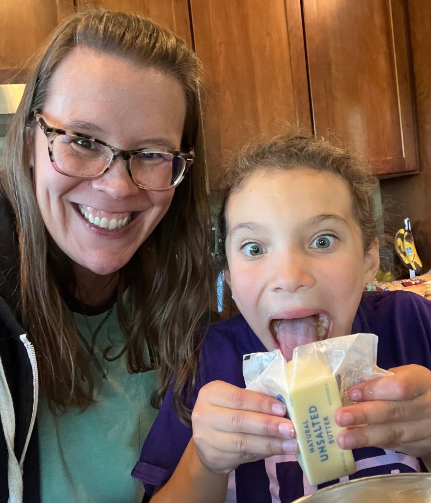An adult female wearing glasses (Keila) is pictured smiling with her arm around her niece who is miming licking a stick of butter. The butter is soft but holding it's shape as it's being tipped into the mixing bowl.
