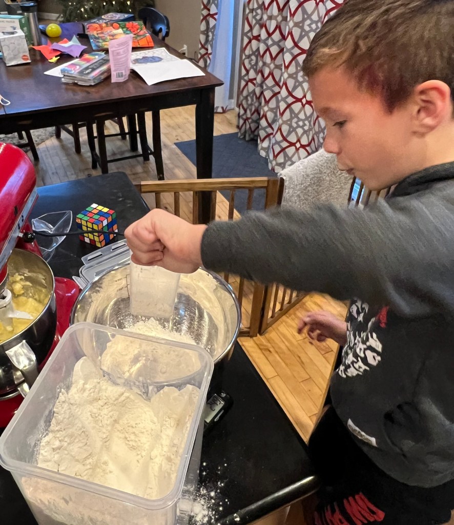 A five year old boy wearing a gray sweatshirt is pictured scooping flour from a tuppereware bin into a bowl resting on a scale. His lips are pursed and there's a Rubix cube in the background.